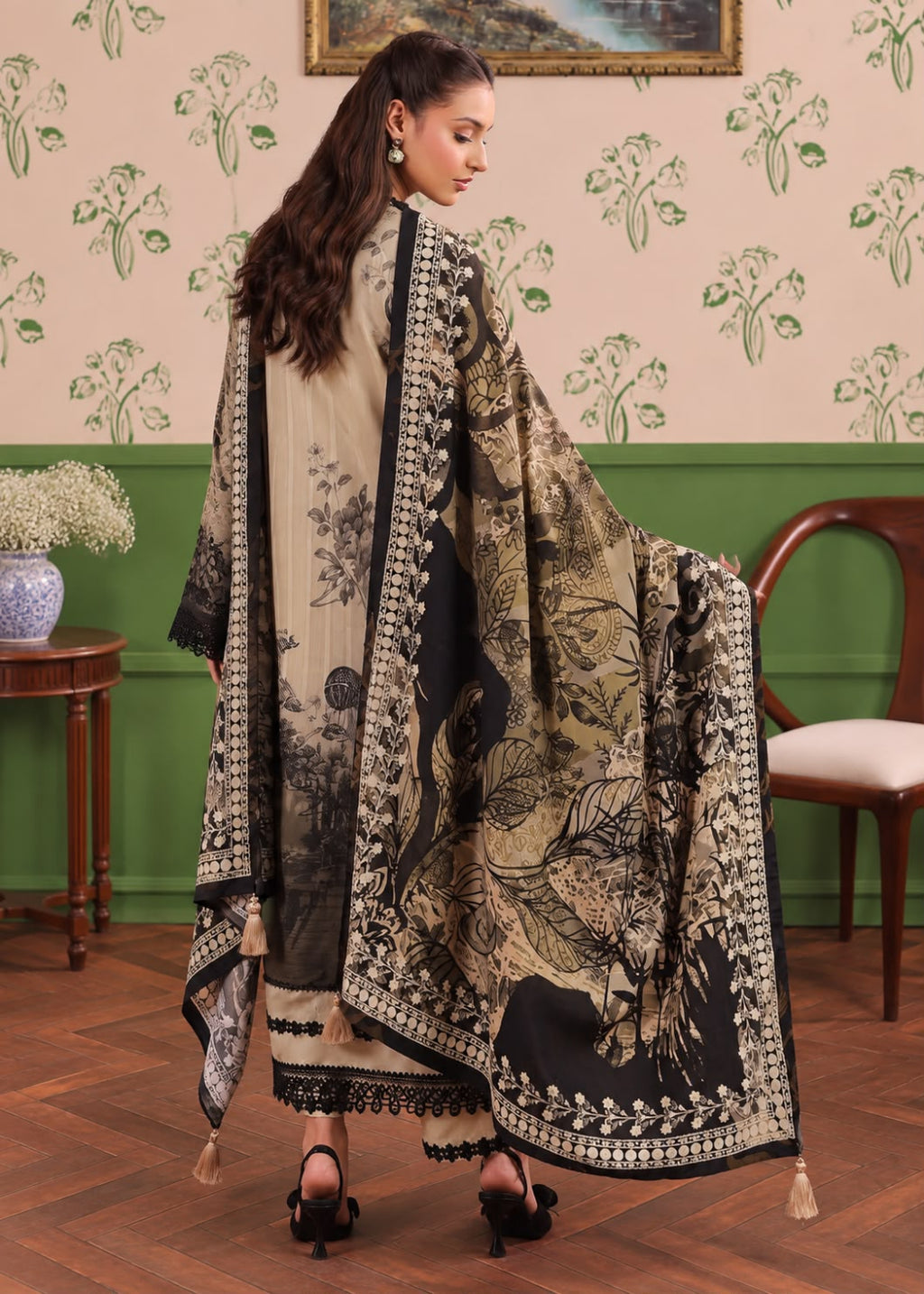 A woman modeling a traditional South Asian outfit with a beige and black printed kurta, matching skirt, and a large matching dupatta with ornate borders, posed in an indoor room with patterned wallpaper and wooden furniture.