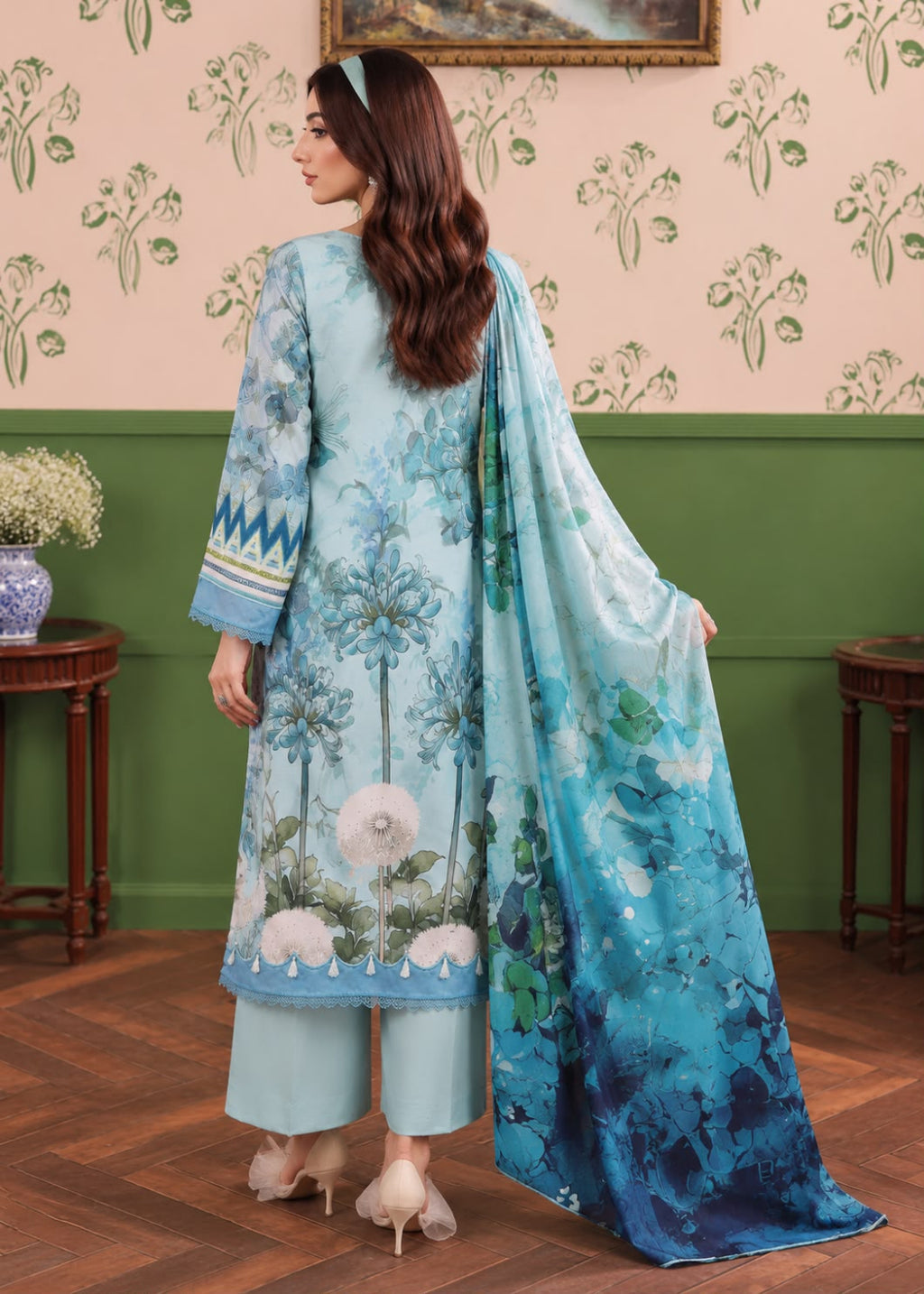 A woman stands with her back to the camera wearing a light blue traditional outfit with a floral print, matching pants, and a long blue dupatta, posed in an indoor room with patterned wallpaper and wooden floors.