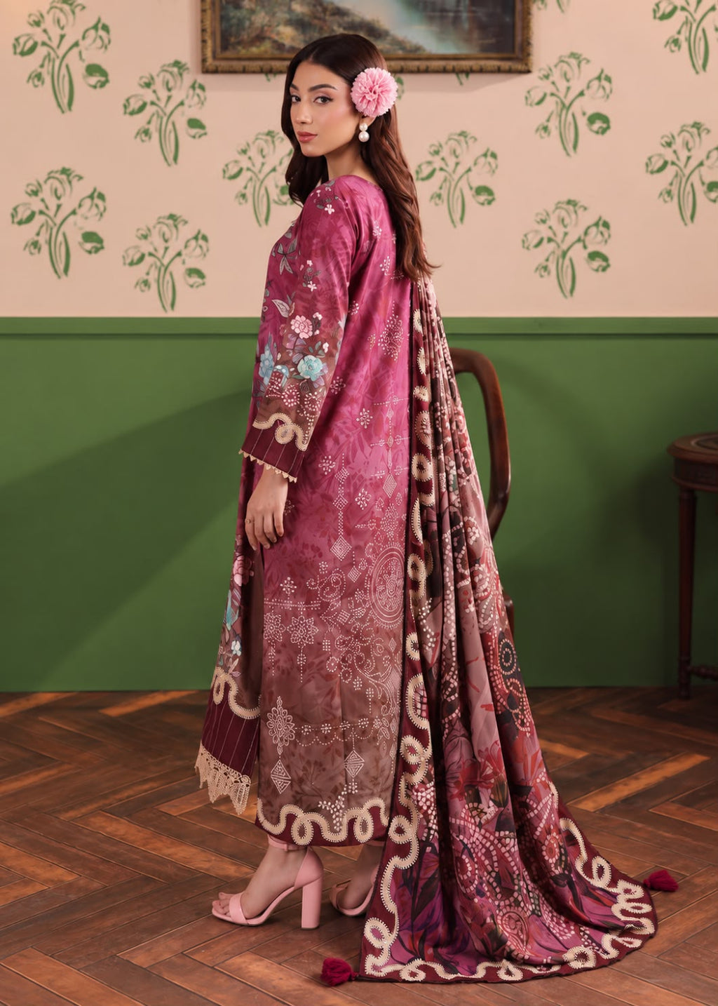 A woman models a traditional three-piece salwar kameez set in a dusty rose/magenta color, featuring intricate floral embroidery, a matching printed dupatta, and a beige lace hem, set against a green and beige patterned wall.
