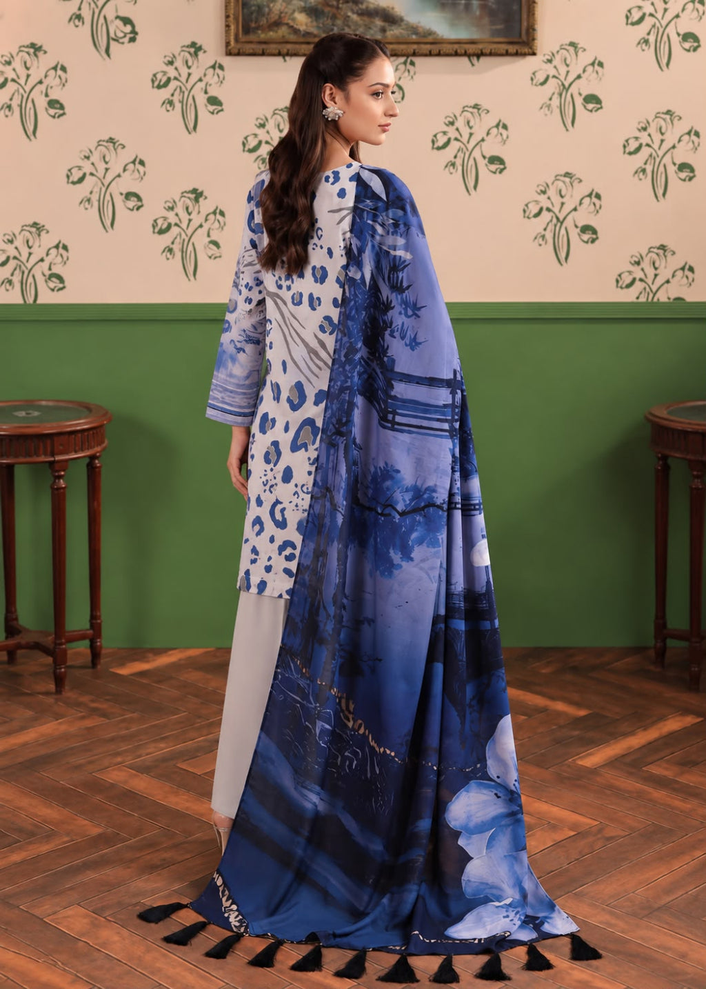 A woman models a traditional South Asian outfit consisting of a white kurta with blue floral patterns, light gray trousers, and a matching blue printed dupatta with black tassels, set against a vintage-style room with green and beige walls.