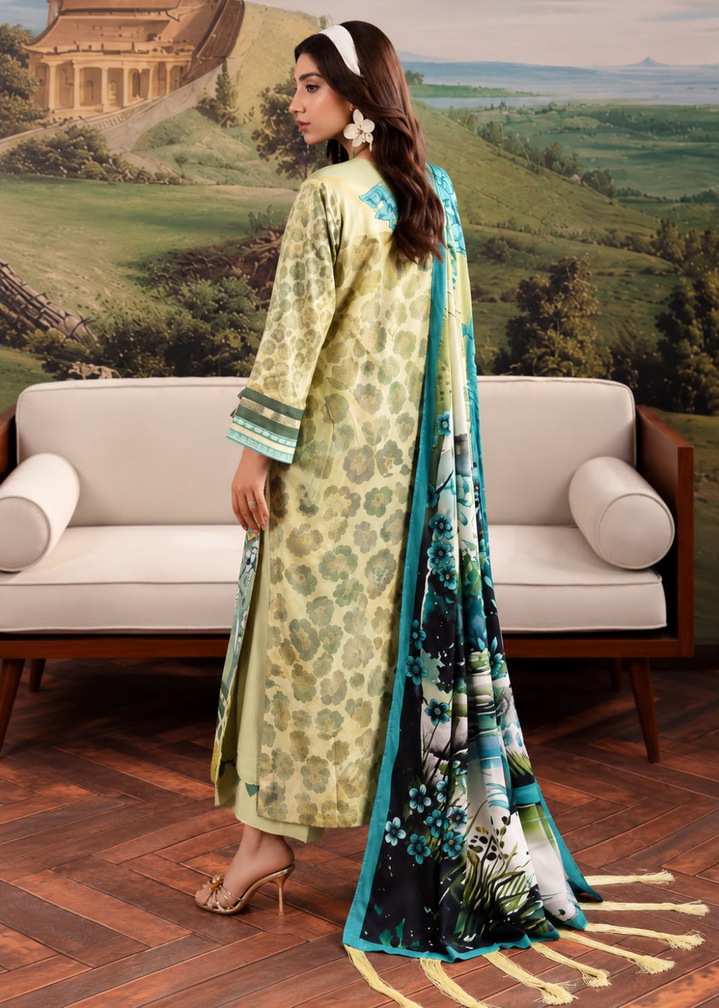 A woman models a traditional South Asian outfit consisting of a light yellow-green patterned kurta, a matching printed dupatta with blue and green floral motifs, and beige heels, posed in front of a cream-colored sofa with a scenic landscape backdrop.