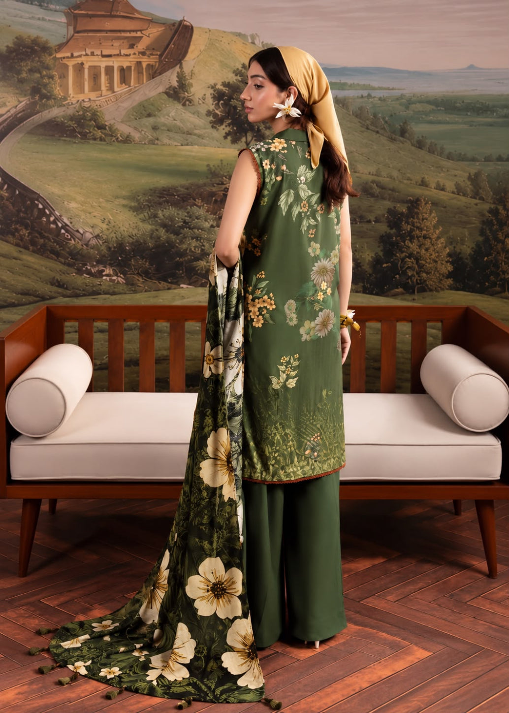 A woman models a three-piece olive green ethnic salwar kameez suit with a floral print, standing in front of a wooden bench and a scenic landscape mural.