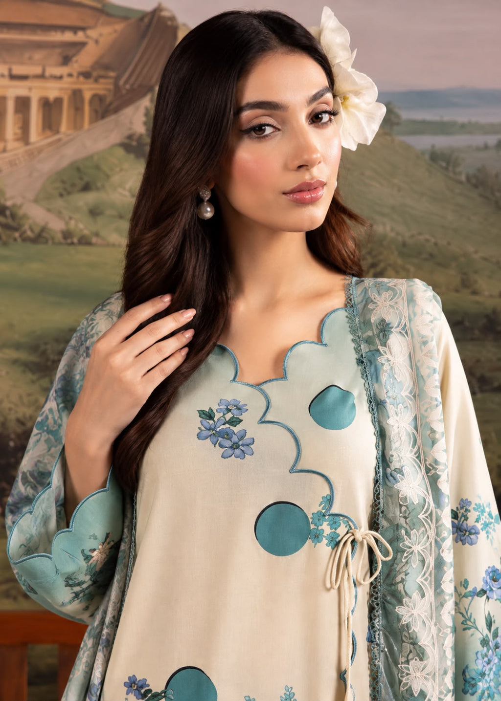 A woman modeling a cream-colored traditional outfit with blue floral embroidery and teal circular cutouts, paired with a matching light blue printed shawl, set against a scenic landscape backdrop.