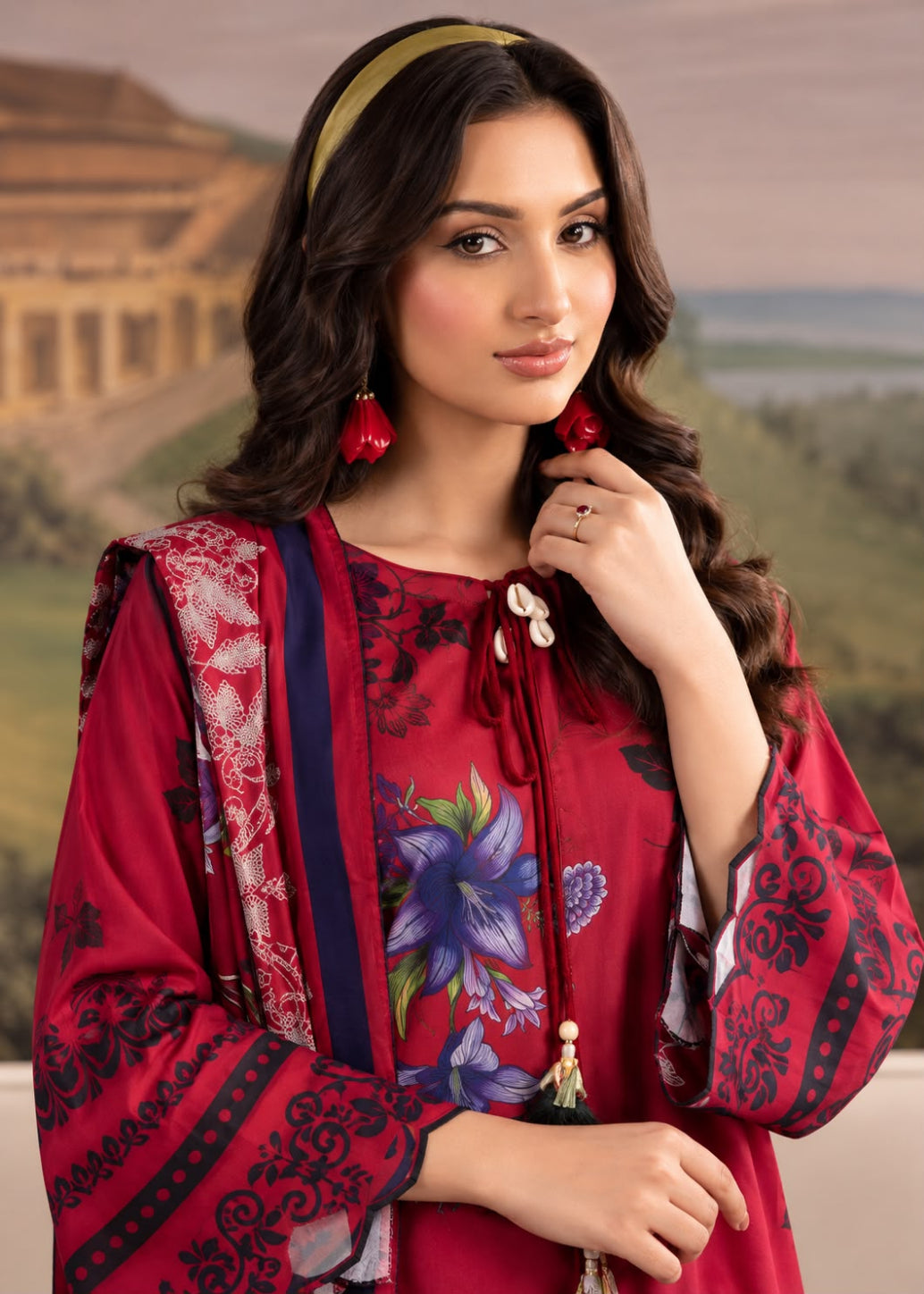 A woman modeling a red printed ethnic kurta set with floral patterns, a matching headband, and red earrings, set against a blurred background of a historic stone structure and green hills.