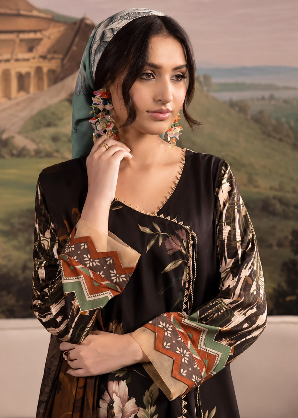 A woman modeling a black printed kurta with multicolor geometric and floral patterns, paired with a patterned headscarf and large floral earrings, set against a scenic landscape backdrop.