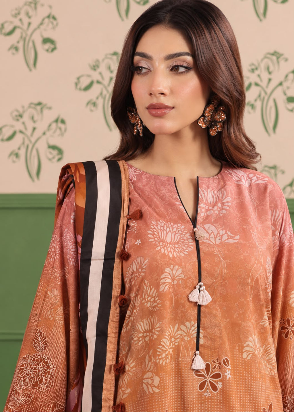 A woman wearing a peach and orange printed salwar suit with a black, white, and orange striped dupatta, set against a patterned cream wall.