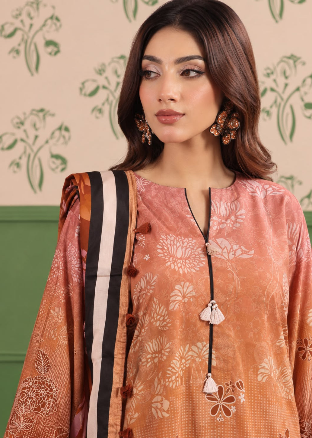 A woman wearing a peach and orange printed salwar suit with a black, white, and orange striped dupatta, set against a patterned cream wall.