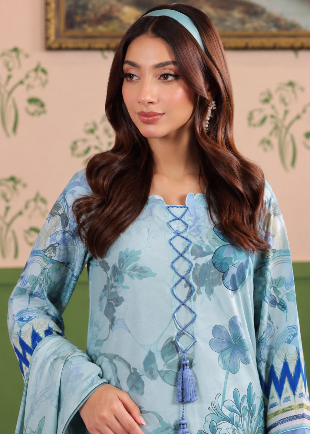 A woman modeling a light blue floral printed traditional outfit with a matching headband, set against a soft beige wall with green floral patterns.