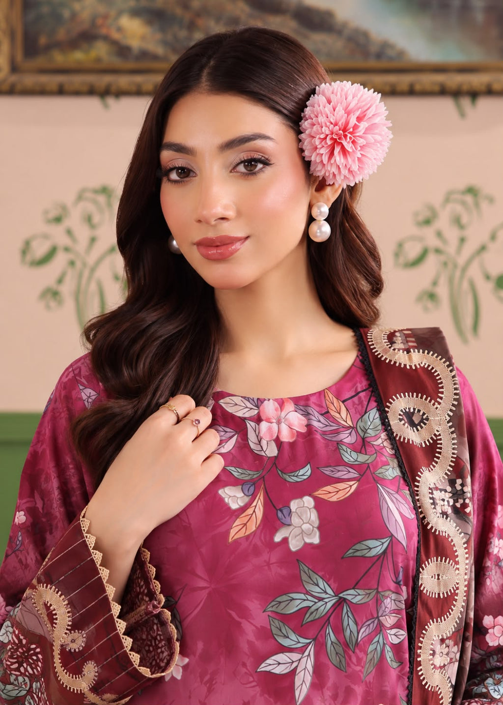 A woman wearing a traditional embroidered outfit in deep magenta with floral patterns and a matching patterned dupatta, styled with a pink flower in her hair.