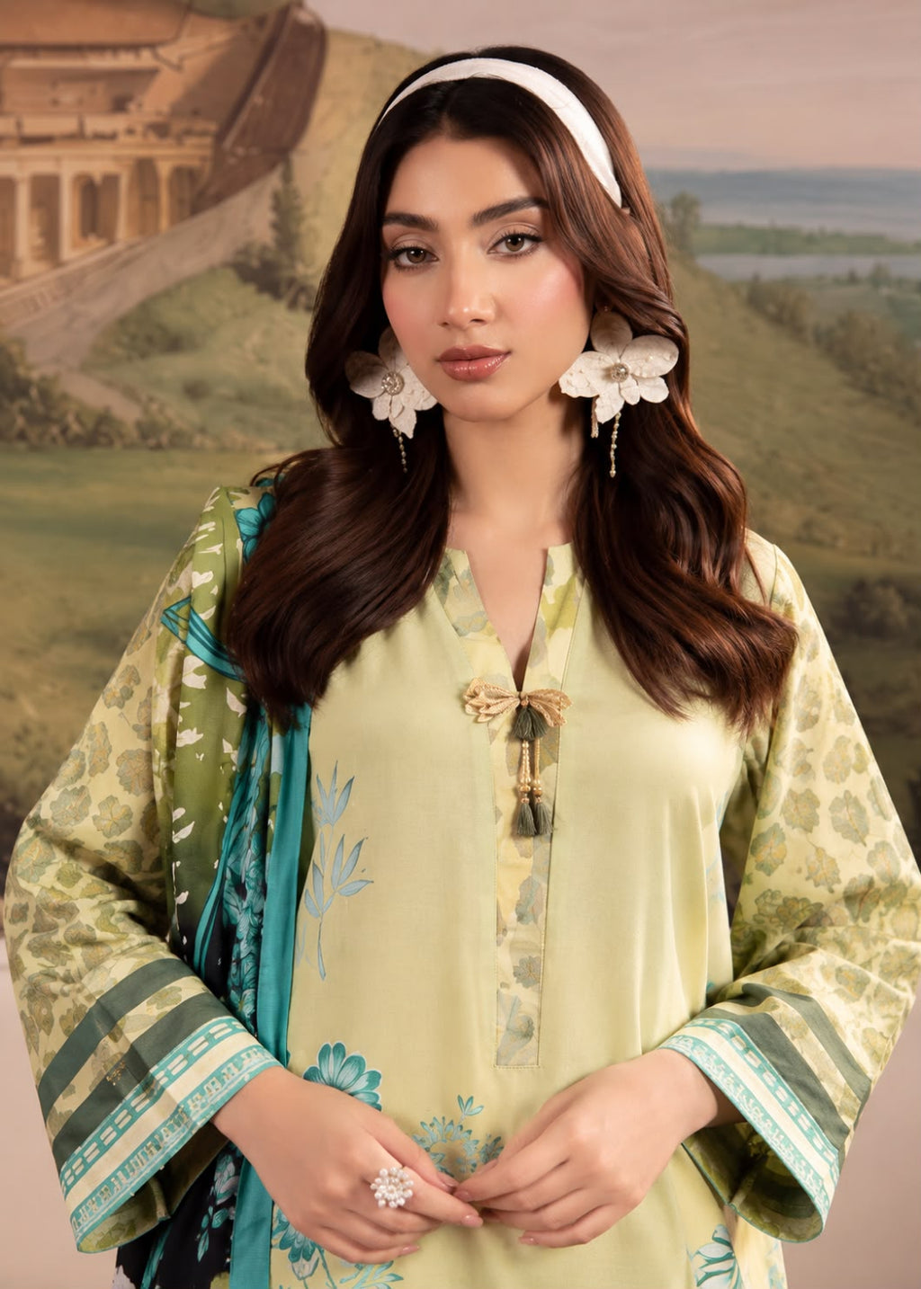 A woman modeling a traditional South Asian outfit featuring a light green kurta with blue floral embroidery, a green patterned dupatta, and a white headband, set against a scenic painted backdrop.