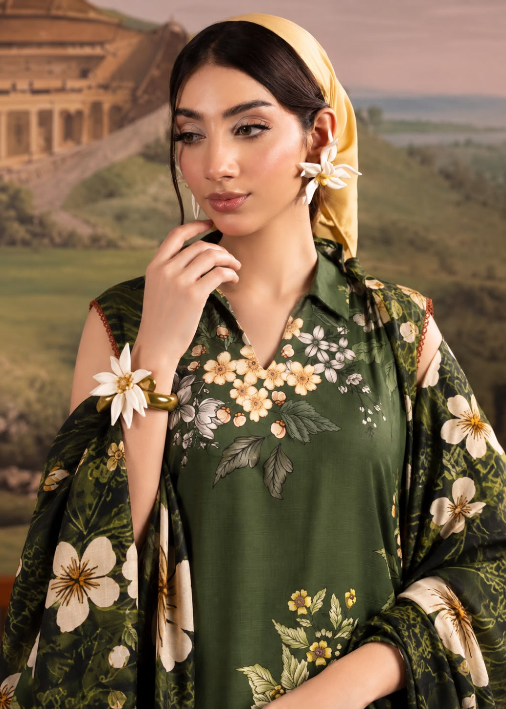 A woman wearing a dark green embroidered traditional outfit with white and yellow floral patterns, a matching yellow headscarf, and white flower earrings, posed against a scenic painted backdrop.