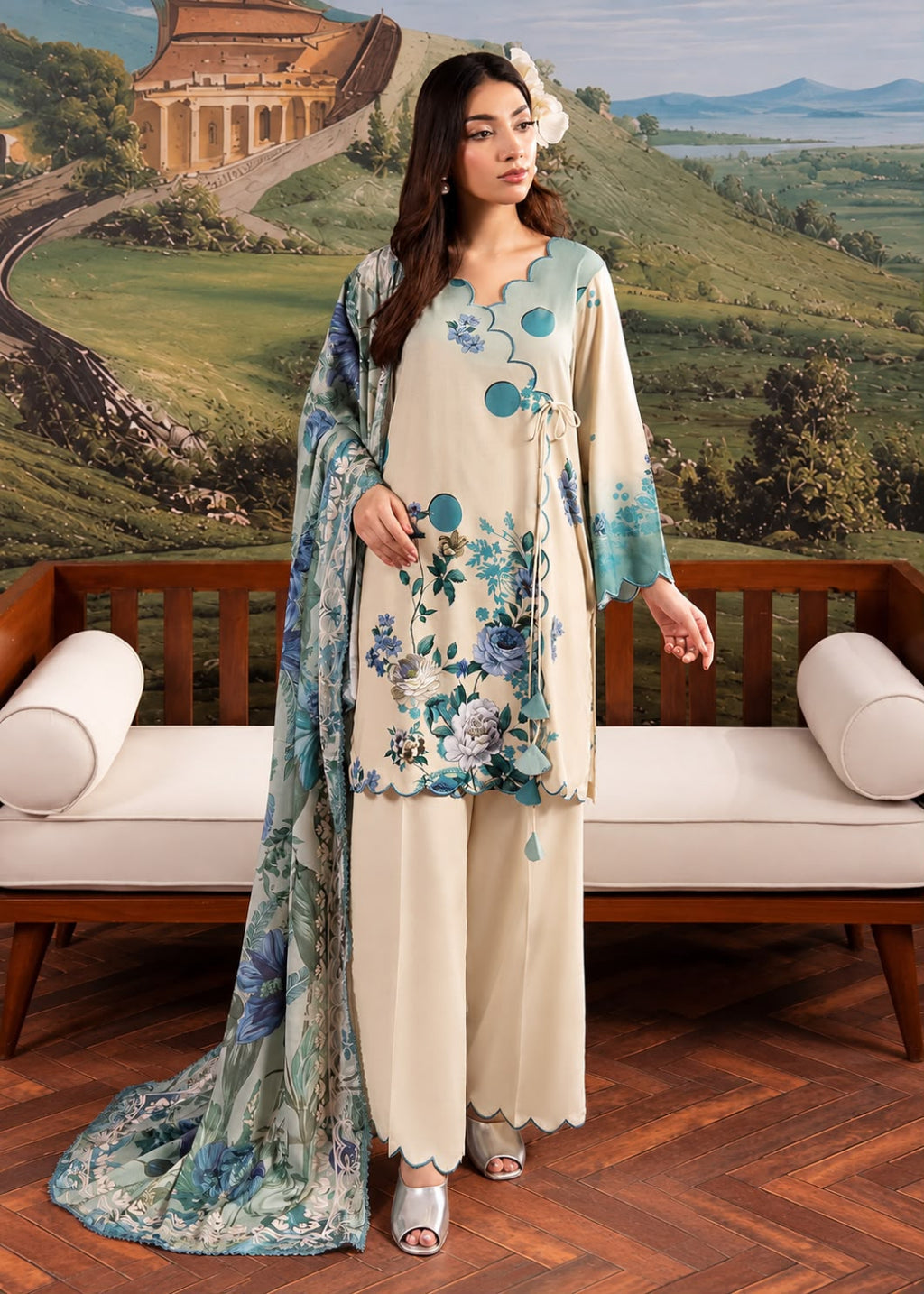 A woman models a traditional South Asian three-piece salwar kameez suit set. The set features a cream-colored kurta with blue and green floral prints, matching wide-leg trousers, and a coordinating printed dupatta.