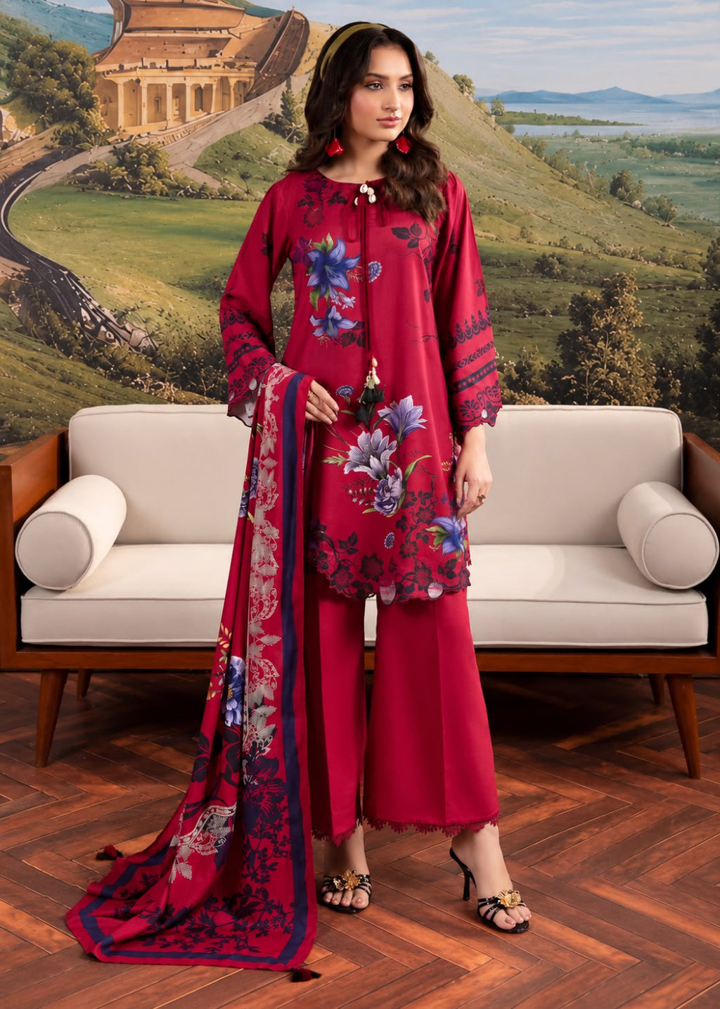A woman models a three-piece salwar kameez suit in deep red with blue and purple floral prints, standing in front of a beige sofa and a scenic landscape backdrop.