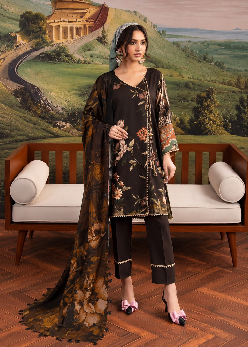 A woman models a three-piece black floral salwar suit set with a matching dupatta, standing in front of a wooden bench and a scenic landscape mural.
