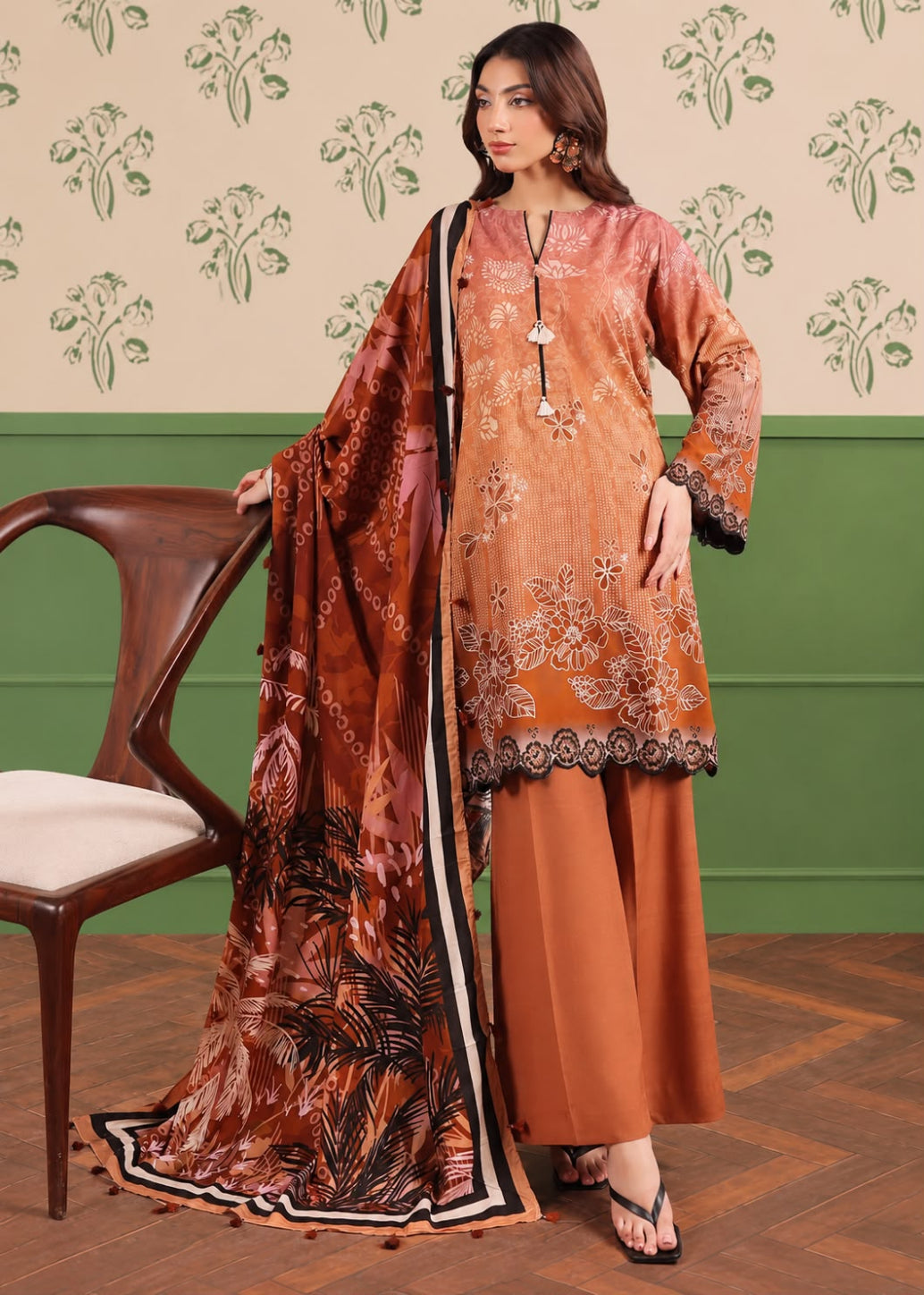 A woman models a traditional three-piece salwar kameez suit in rust orange and brown, featuring floral embroidery on the kurta, wide-leg trousers, and a matching printed dupatta, set against a patterned wallpaper and wooden chair background.