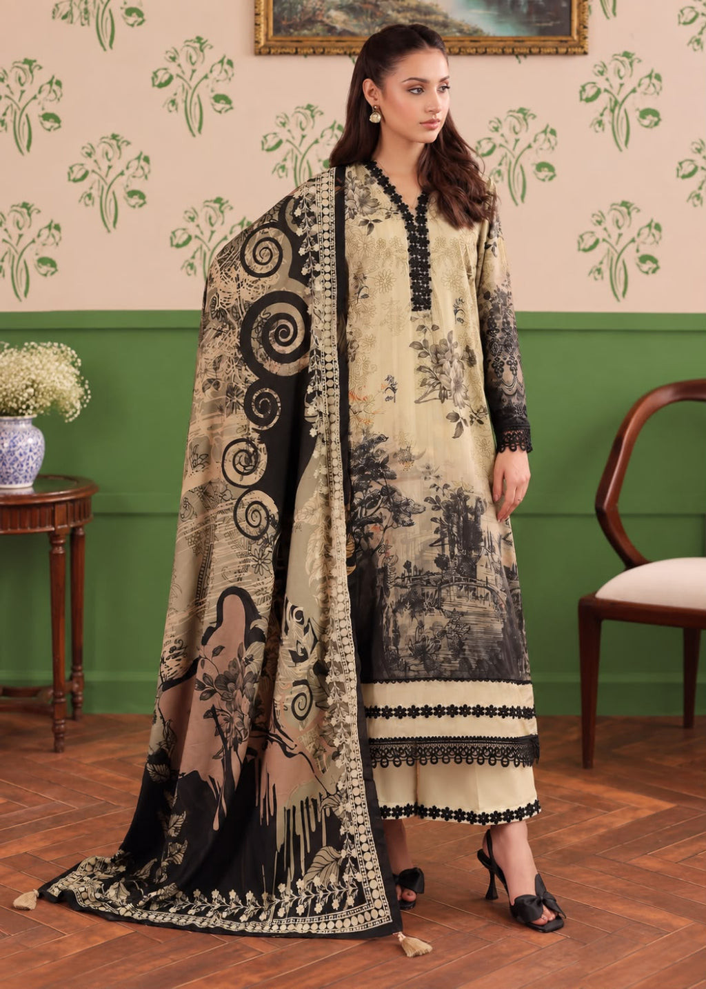 A woman models a traditional South Asian salwar kameez suit with a beige and black landscape print, featuring a matching printed dupatta, in an indoor room with green and beige walls.