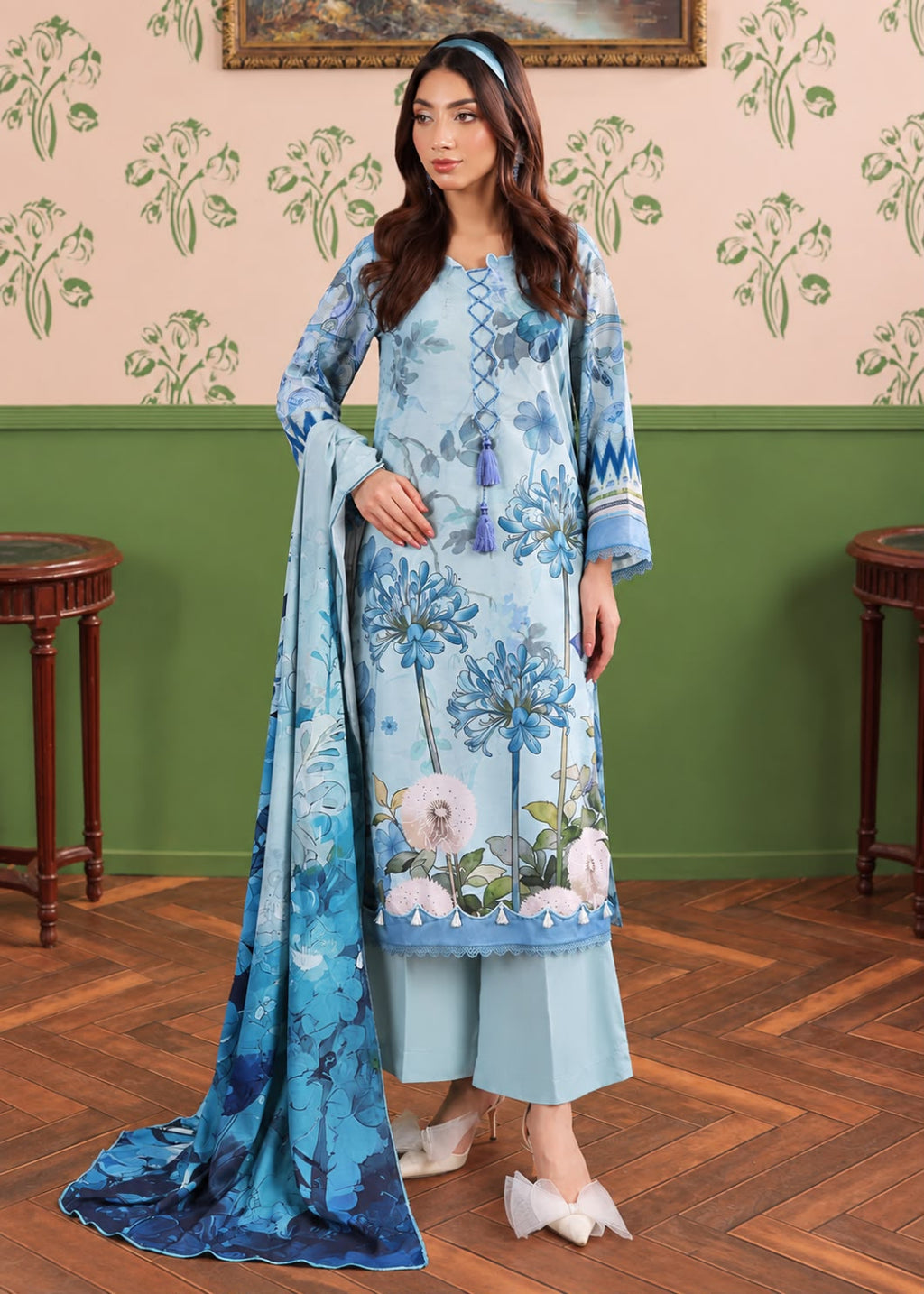A woman models a light blue three-piece traditional South Asian outfit with floral embroidery, a matching dupatta, and wide-leg pants in a decorated room with patterned wallpaper and wooden floors.
