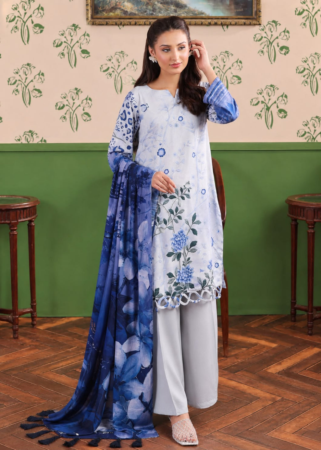 A woman models a traditional South Asian salwar kameez set featuring a white kameez with blue floral prints, light gray salwar pants, and a blue floral dupatta, set against a green and beige patterned wall.