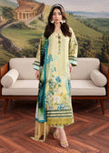 A woman models a traditional three-piece salwar kameez suit in a pale yellow-green color, featuring a floral print, a matching dupatta, and a black embroidered border. She stands in front of a beige sofa with a scenic landscape backdrop.