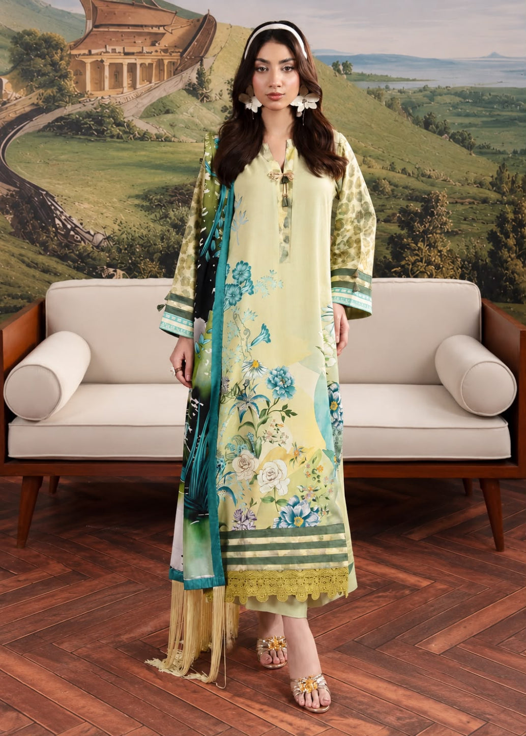 A woman models a traditional three-piece salwar kameez suit in a pale yellow-green color, featuring a floral print, a matching dupatta, and a black embroidered border. She stands in front of a beige sofa with a scenic landscape backdrop.