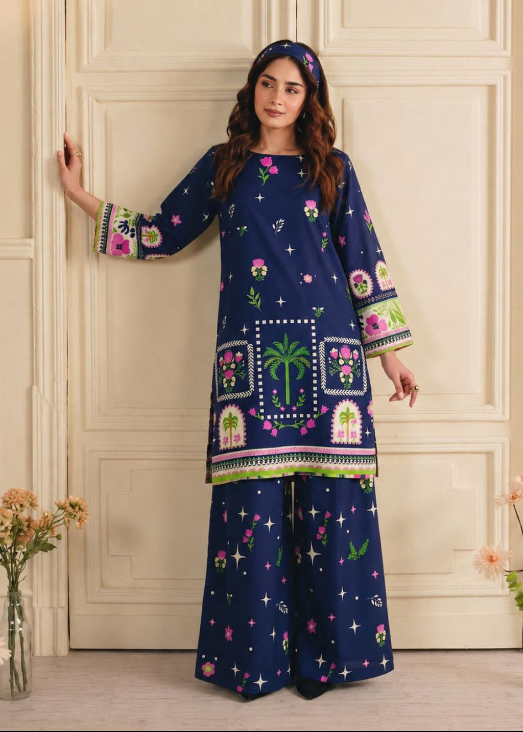 A woman models a navy blue three-piece ethnic suit with colorful floral and geometric prints, standing in front of a cream-colored paneled door.