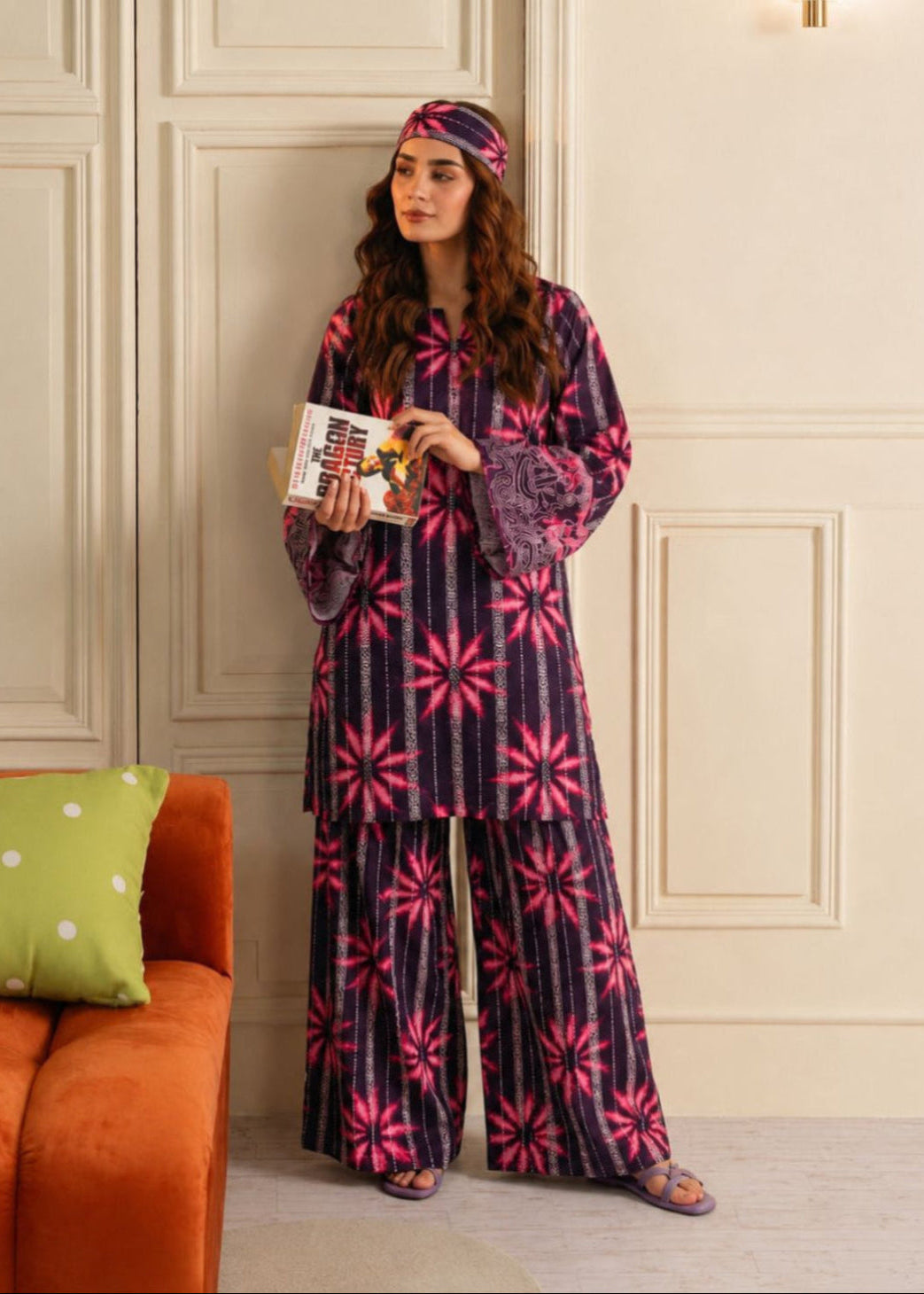 A woman stands indoors wearing a matching two-piece pajama set with a dark purple base and bright pink floral starburst patterns. She is holding a book and wearing a matching headband.
