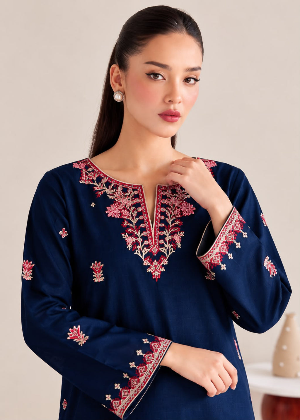 A woman wearing a navy blue long-sleeve kurta with intricate red, pink, and gold floral embroidery around the neckline and cuffs, posed against a neutral beige background.