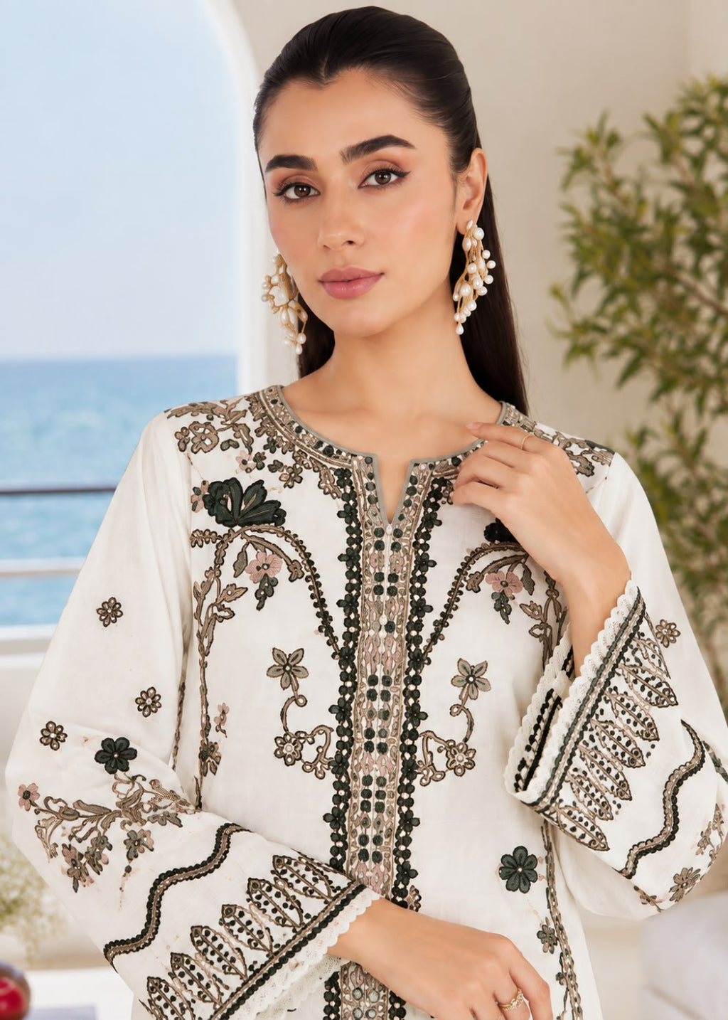 A woman wearing a white embroidered tunic with intricate black and beige floral patterns, styled with pearl-like drop earrings, set against a soft-focus background of a balcony and greenery.
