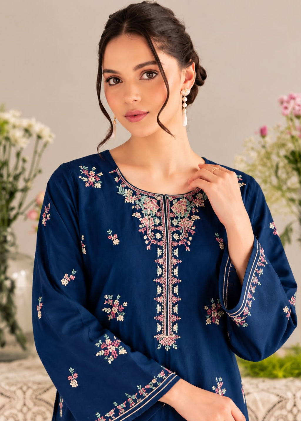 A woman wearing a navy blue embroidered kurta with floral patterns and a round neckline, posed against a soft beige background with blurred flowers.