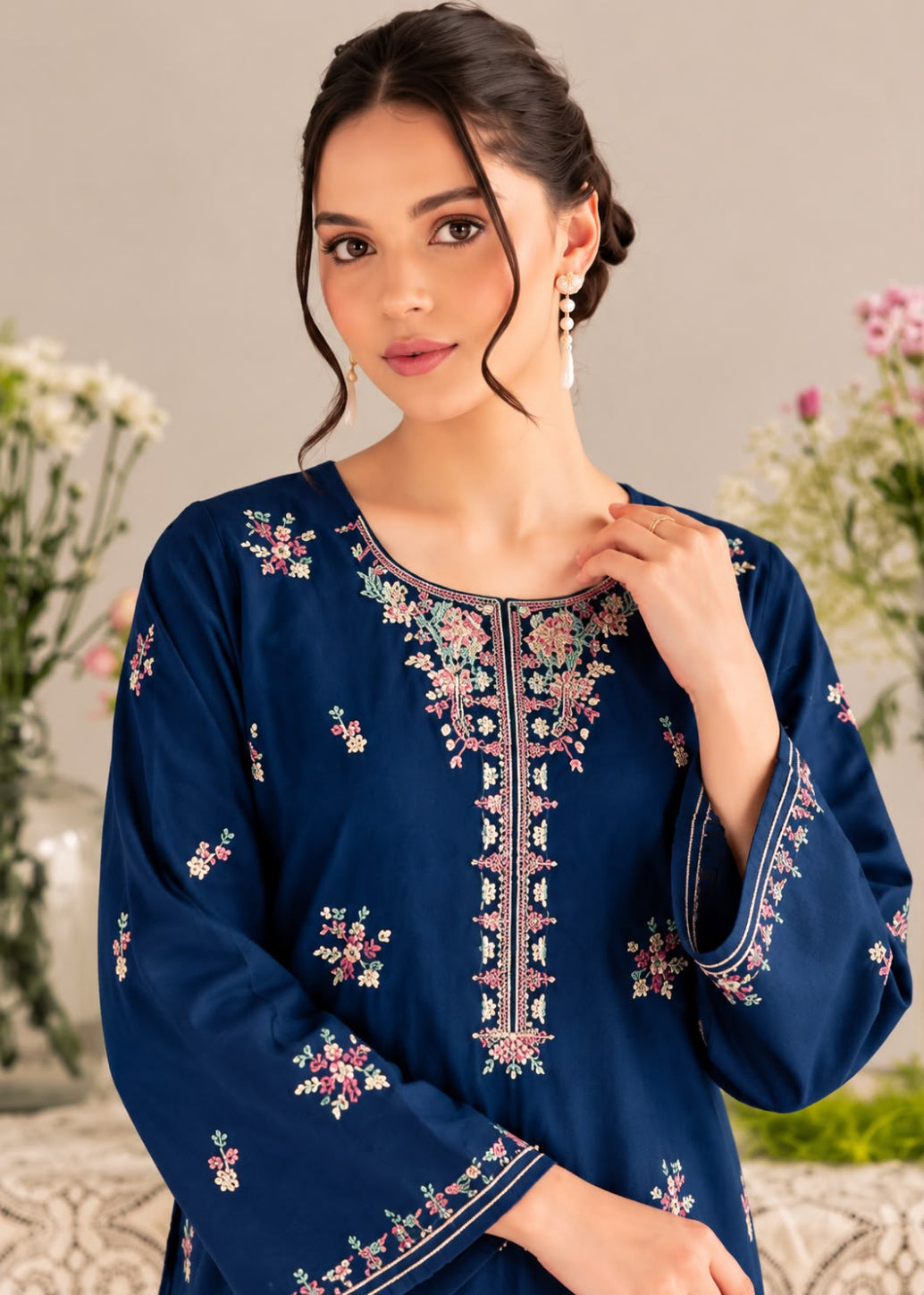A woman wearing a navy blue embroidered kurta with floral patterns and a round neckline, posed against a soft beige background with blurred flowers.