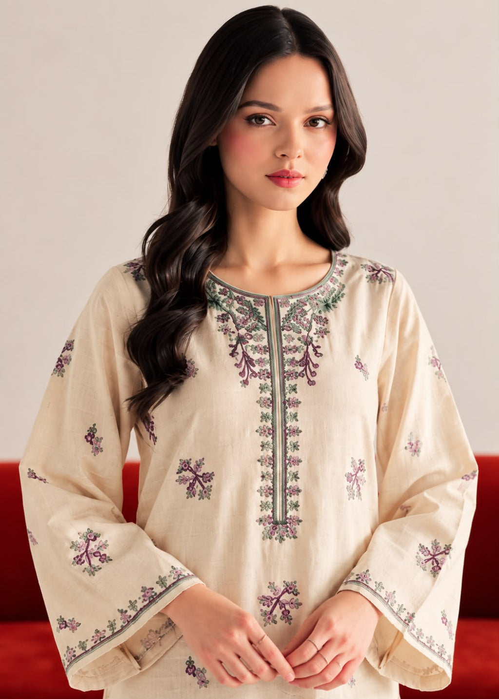 A woman wearing a cream-colored embroidered tunic top with purple and green floral patterns, posed against a neutral background with a red sofa visible behind her.