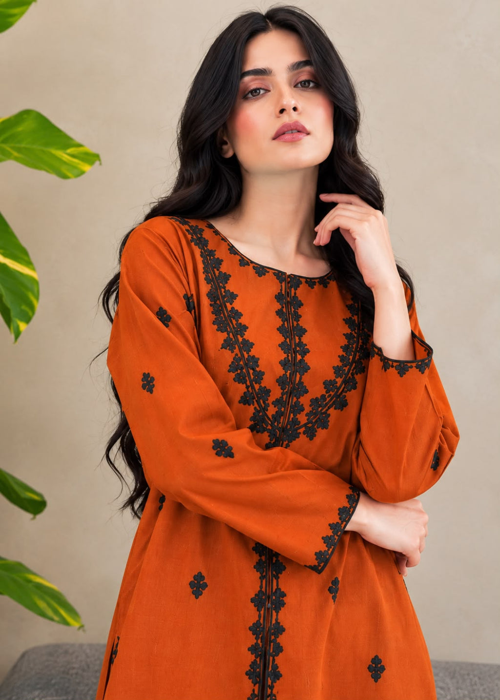 A woman wearing a burnt orange long-sleeve kurta with black floral embroidery, posed against a neutral beige wall with green leaves on the side.