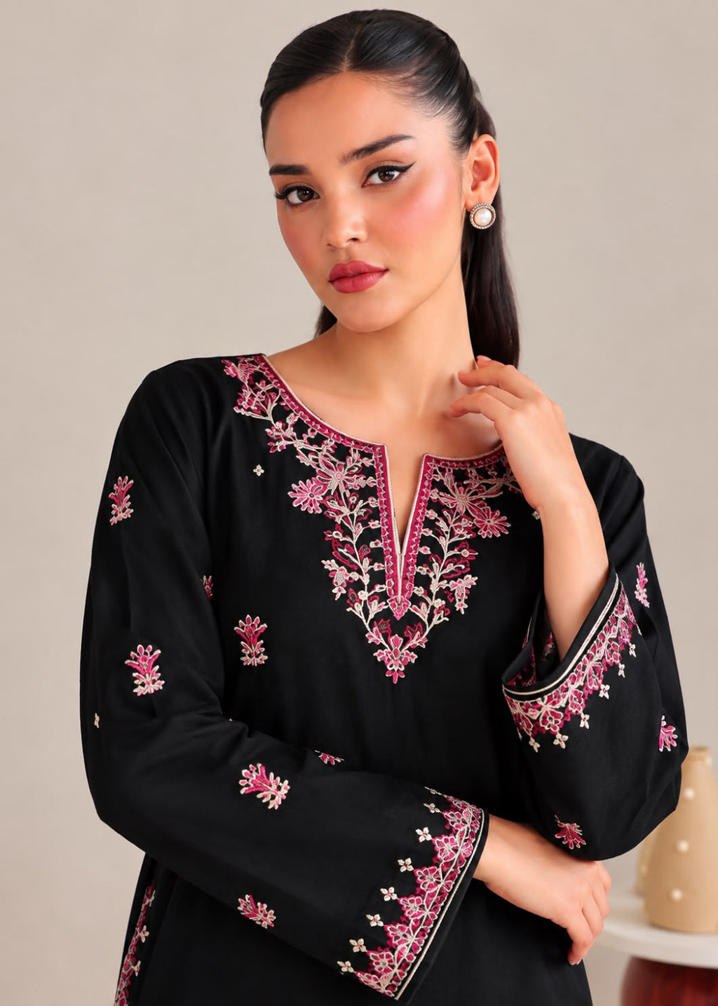 A woman modeling a black long-sleeve kurta with intricate pink and white floral embroidery around the split neckline and cuffs, set against a neutral beige background.