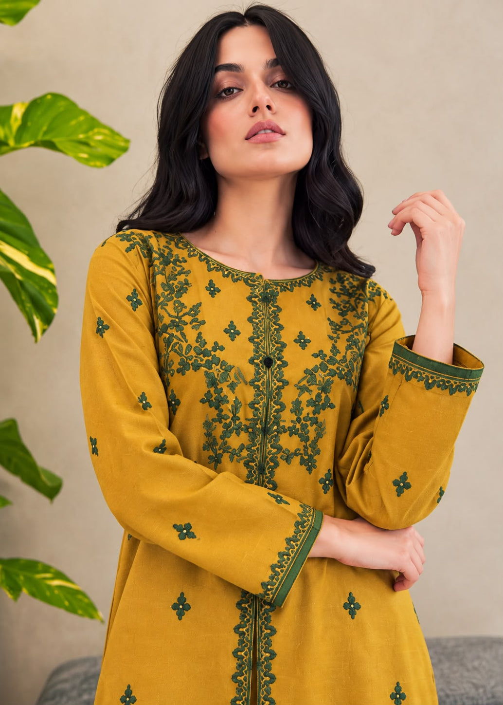 A woman wearing a mustard yellow long kurta with dark green floral embroidery, posed against a neutral beige wall with green leaves on the side.