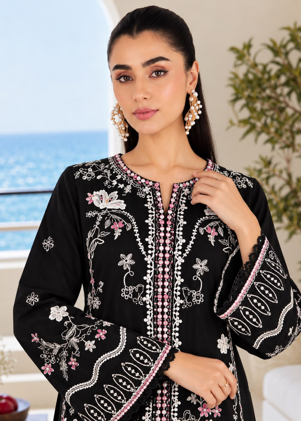 A woman wearing a black embroidered tunic with white and pink floral patterns and beadwork, posed indoors with a window showing the ocean in the background.