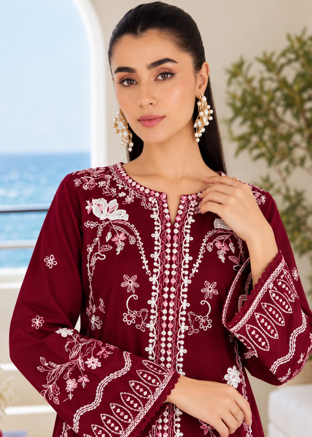A woman wearing a deep maroon embroidered tunic with intricate white floral and geometric patterns, styled with pearl-like drop earrings, set against a softly blurred background of a balcony and the sea.