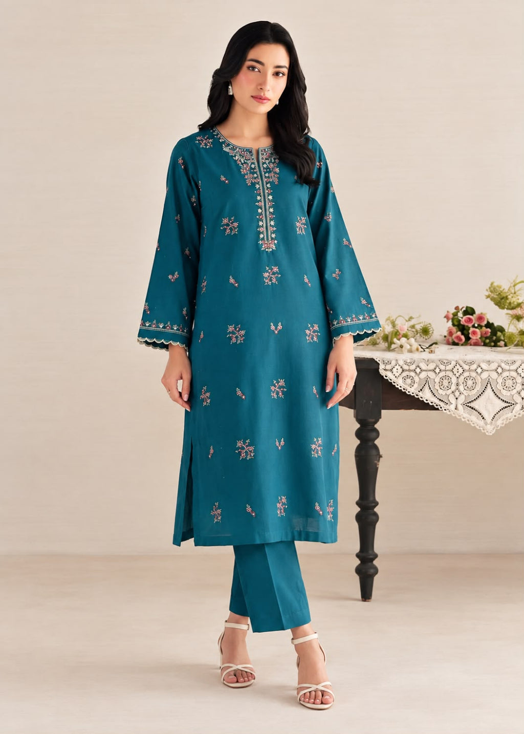 A woman modeling a teal blue-green kurta set with silver embroidery, paired with matching trousers and white heeled sandals, standing in a studio setting with a decorative table in the background.