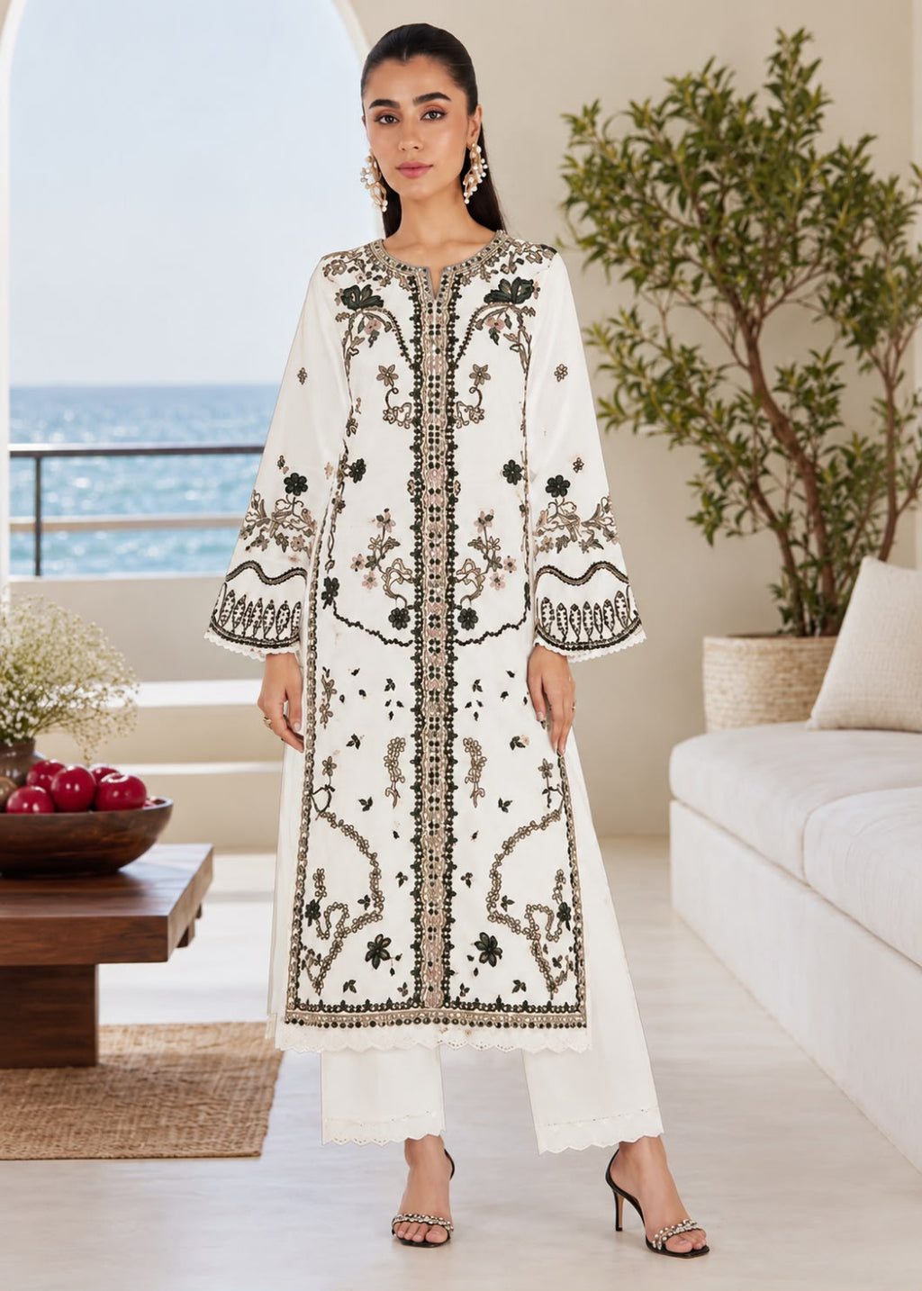 A woman models a long white embroidered kurta with black and gold detailing, paired with matching white trousers, in a bright, modern living room with a view of the ocean in the background.
