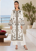A woman models a long white embroidered kurta with black and gold detailing, paired with matching white trousers, in a bright, modern living room with a view of the ocean in the background.