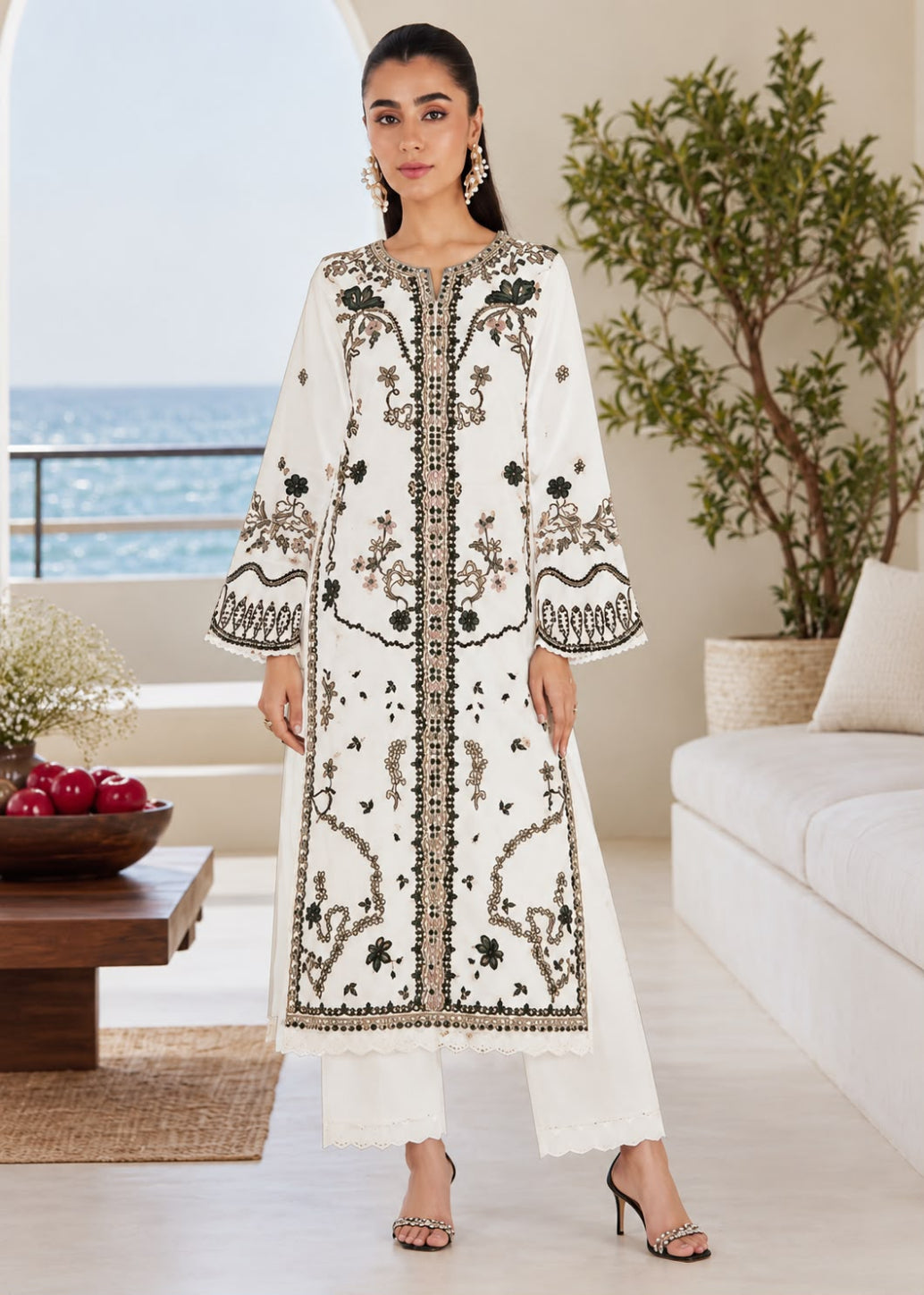A woman models a long white embroidered kurta with black and gold detailing, paired with matching white trousers, in a bright, modern living room with a view of the ocean in the background.