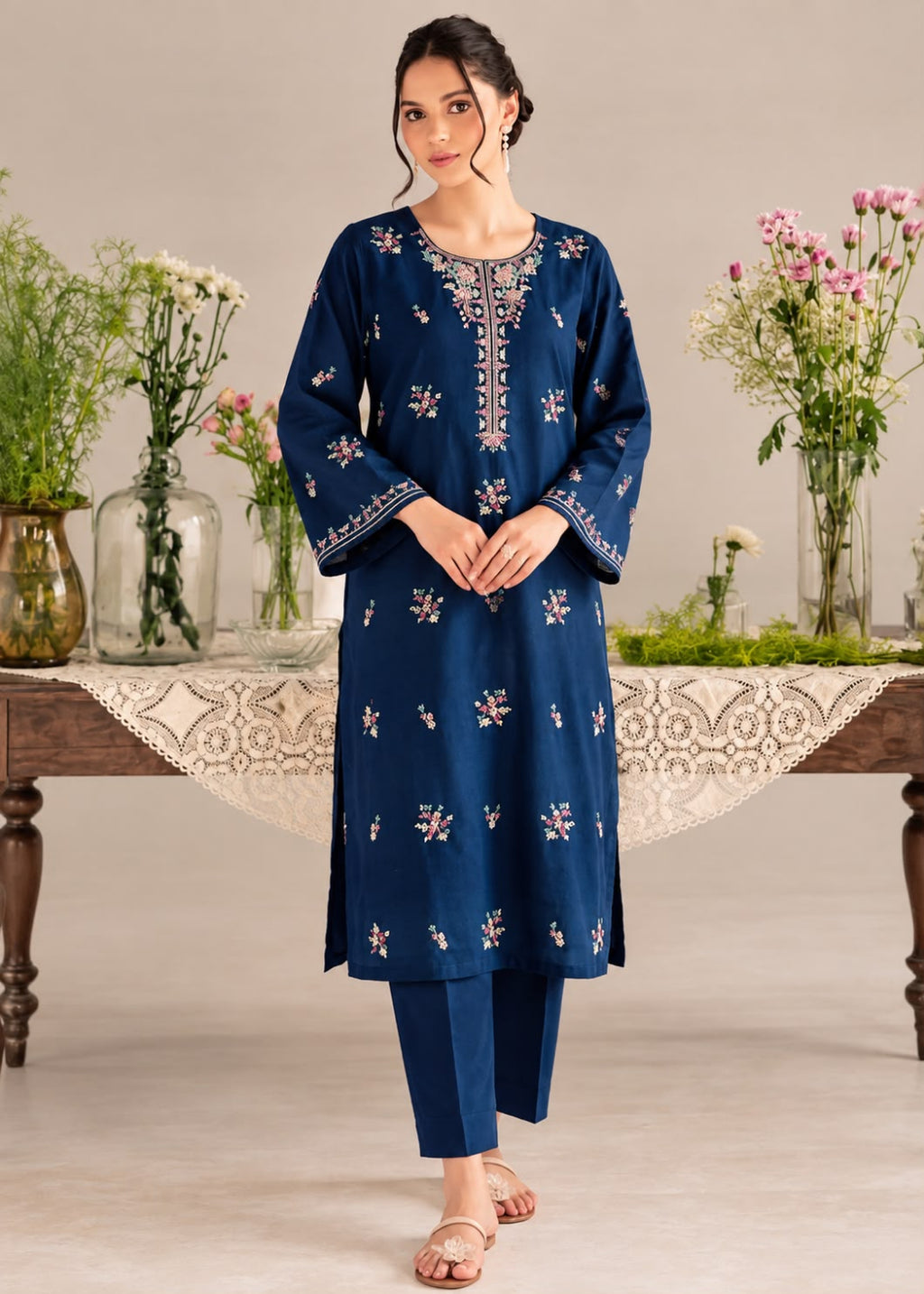 A woman models a navy blue three-piece Indian ethnic suit with floral embroidery, consisting of a long kurta, matching trousers, and a white embroidered inner neck scarf, set against a neutral studio background with decorative floral arrangements.