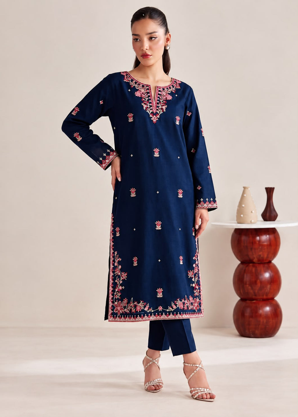 A woman models a navy blue long kurta with pink and gold embroidery, paired with matching navy pants and clear high-heeled sandals, set against a neutral studio background with decorative vases.