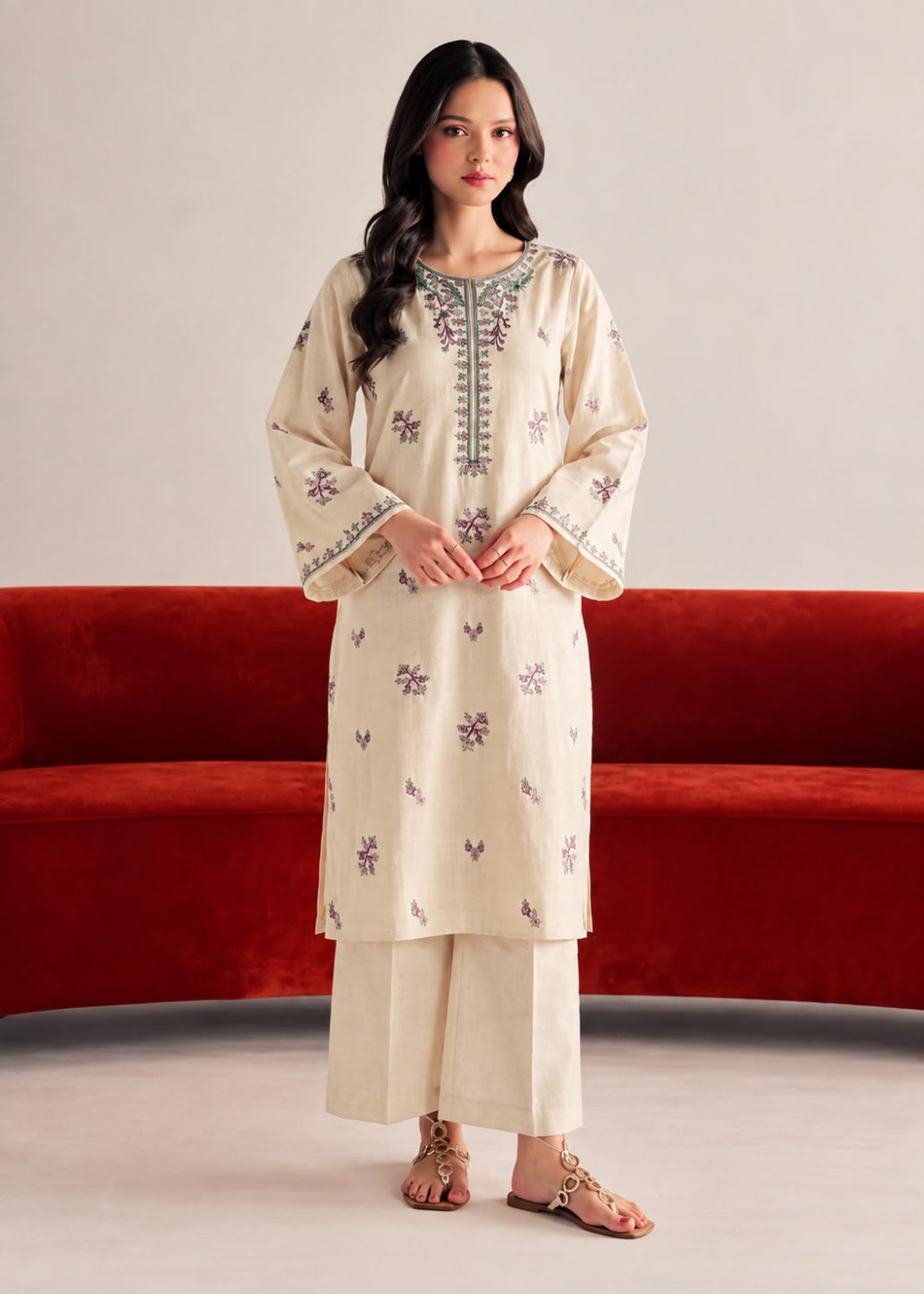 A woman modeling a cream-colored Indian ethnic kurta and pant set with purple floral embroidery and silver mirror work on the neckline, standing in front of a red velvet sofa.