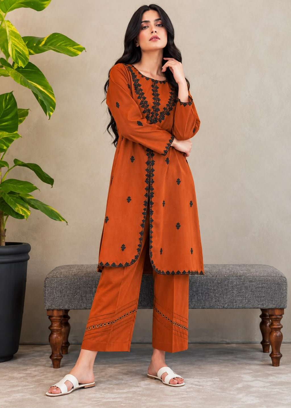 A woman models a rust-orange kurta and trouser set with black embroidered details, standing in front of a gray bench and a potted plant.