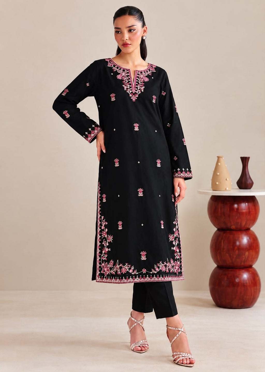 A woman models a black long kurta with pink floral embroidery and silver mirror work details, paired with black pants and silver heels, in a studio setting.