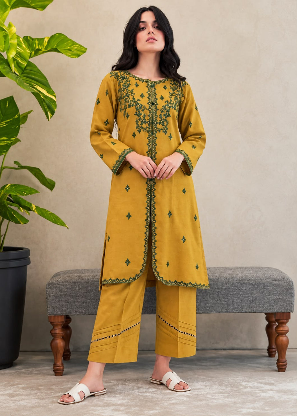 A woman models a mustard yellow kurta and trouser set with green embroidery, standing in front of a gray bench and a potted plant.