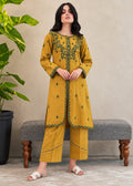 A woman models a mustard yellow kurta and trouser set with green embroidery, standing in front of a gray bench and a potted plant.