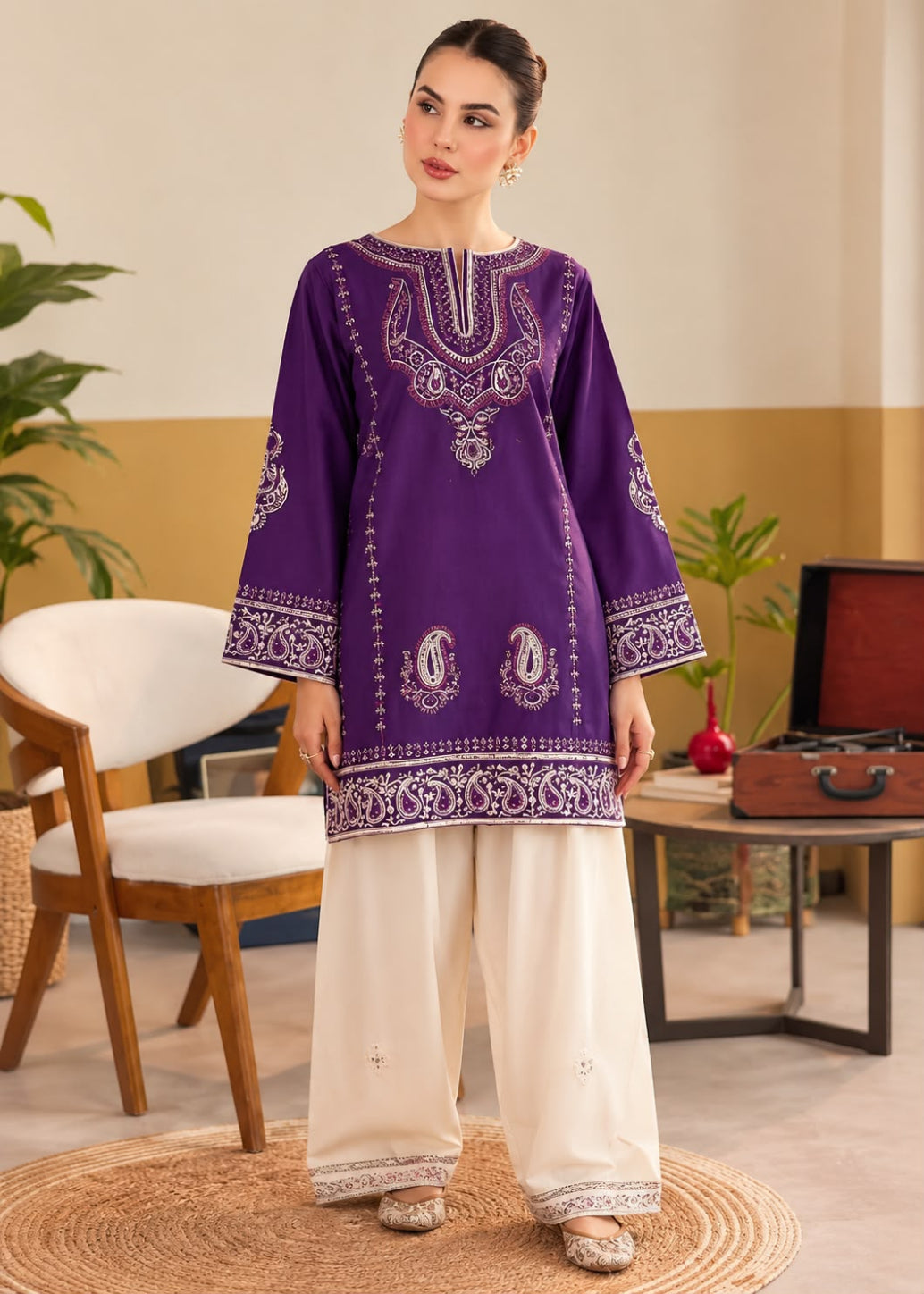 A woman modeling a purple embroidered kurta with white wide-leg pants in a styled indoor setting with a chair, table, and plants.