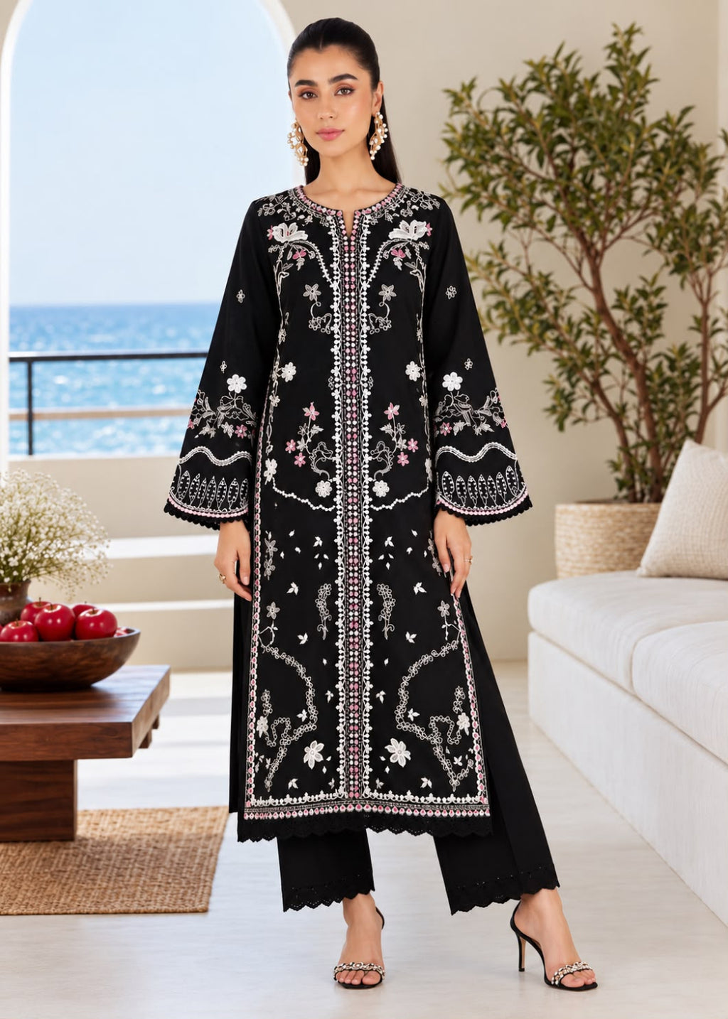 A woman stands in a bright living room wearing a long black embroidered kurta with wide sleeves, scalloped edges, and white floral and geometric detailing. She is paired with black pants and metallic heeled sandals, with a sofa, table, and ocean view in the background.