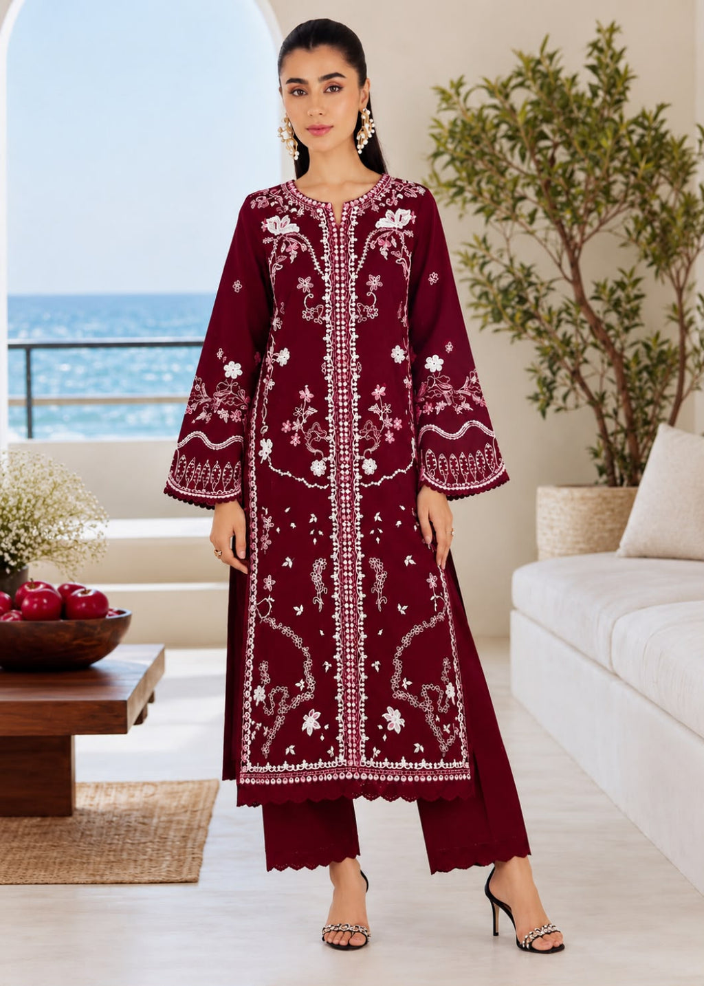 A woman models a deep maroon three-piece kurta set with intricate white floral embroidery, standing in a bright, modern living room with a view of the ocean in the background.